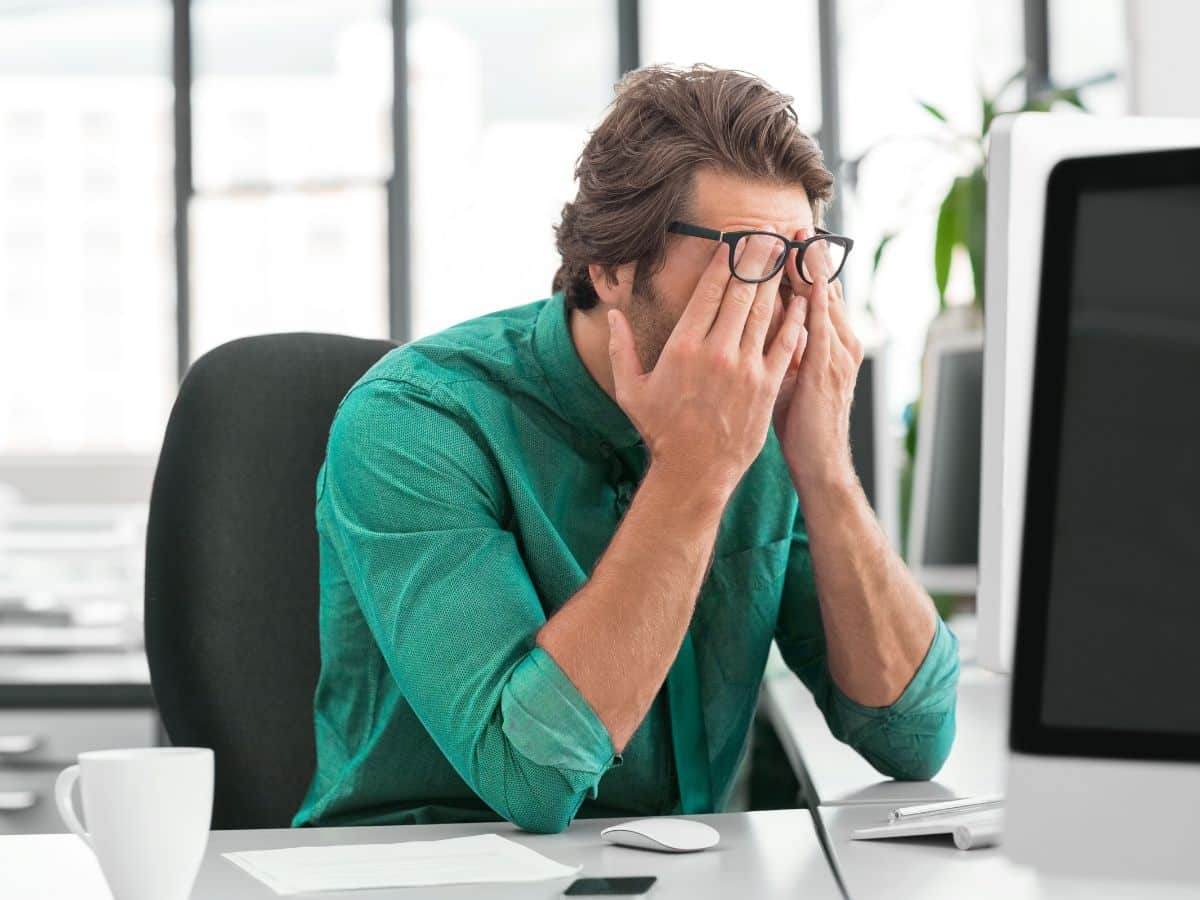 a man sits at his desk, stressed, with his hands on his face, pushing his glasses up; he is wearing a netgain-green button down shirt