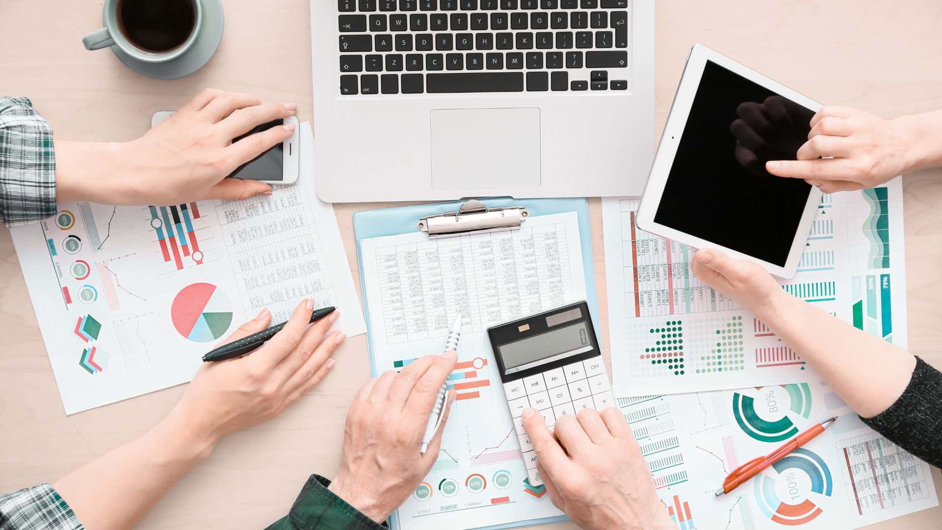 overhead view of a desk, three people's hands hold various papers, pens, and a calculator amid a conversation seemingly about finances