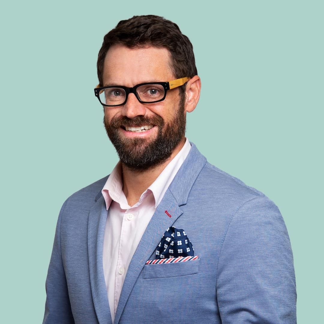 Eric Strawbridge; dark brown-haired man, smiling, wearing glasses, he has on a light blue blazer and light pink button-up, with a cheery patterned pocket-square