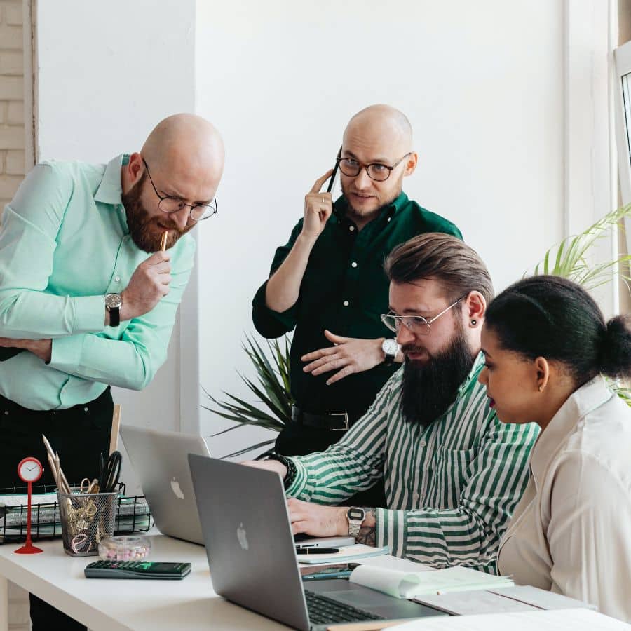 two people standing next to a desk at which two people sit, they are conversing over two computers, one person standing is on the phone