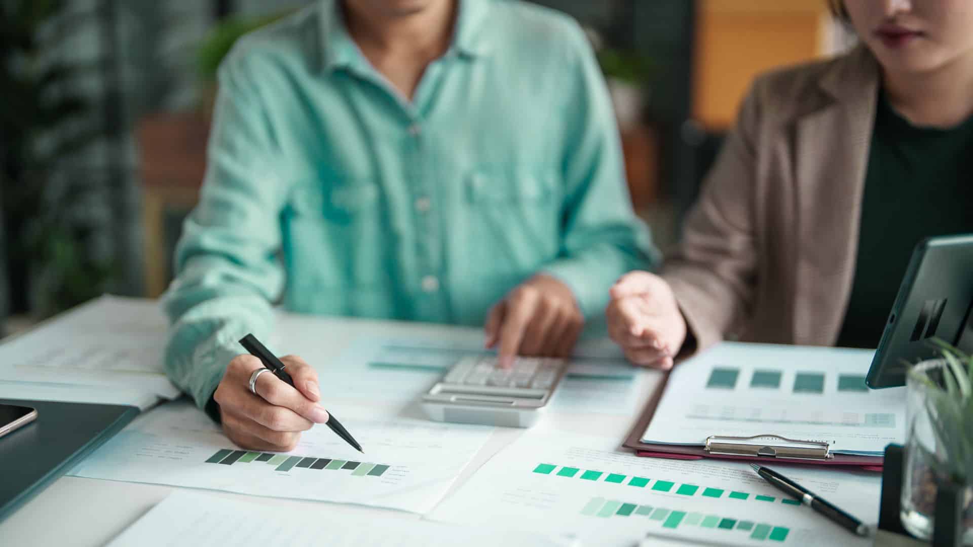 two people sitting at a desk, looking over financial charts, their faces are not visible