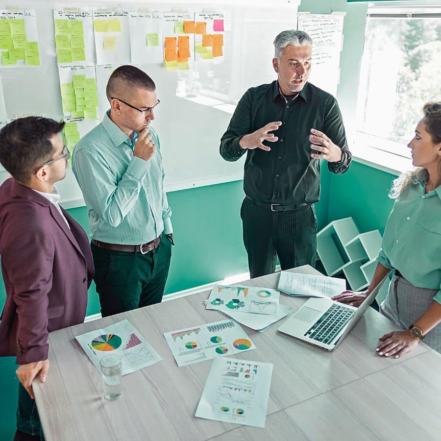 group of business professionals having a conversation, one sits at a desk while the others stand around the end of it, discussing financial charts on paper