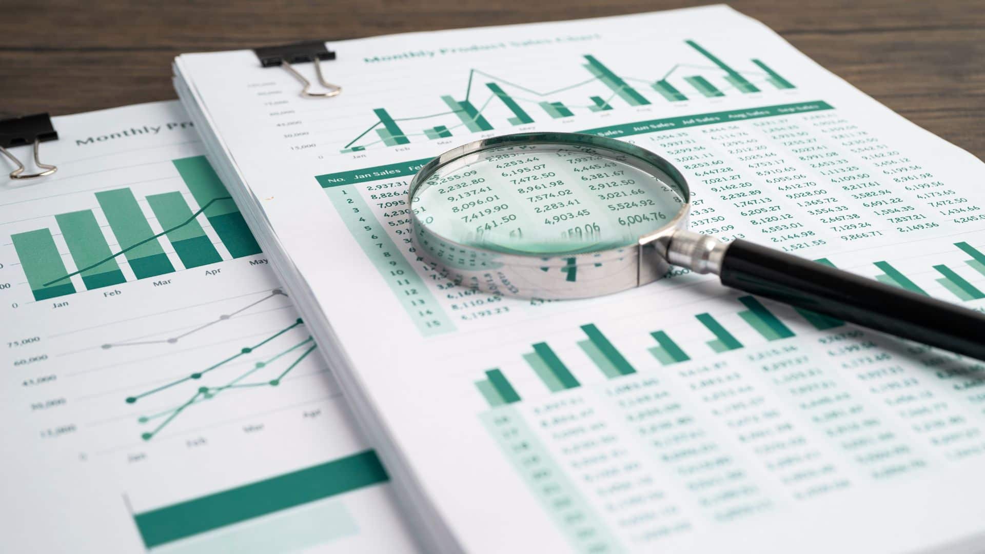 stack of papers in binder clips with financial charts on them; on the bundle sits a magnifying glass
