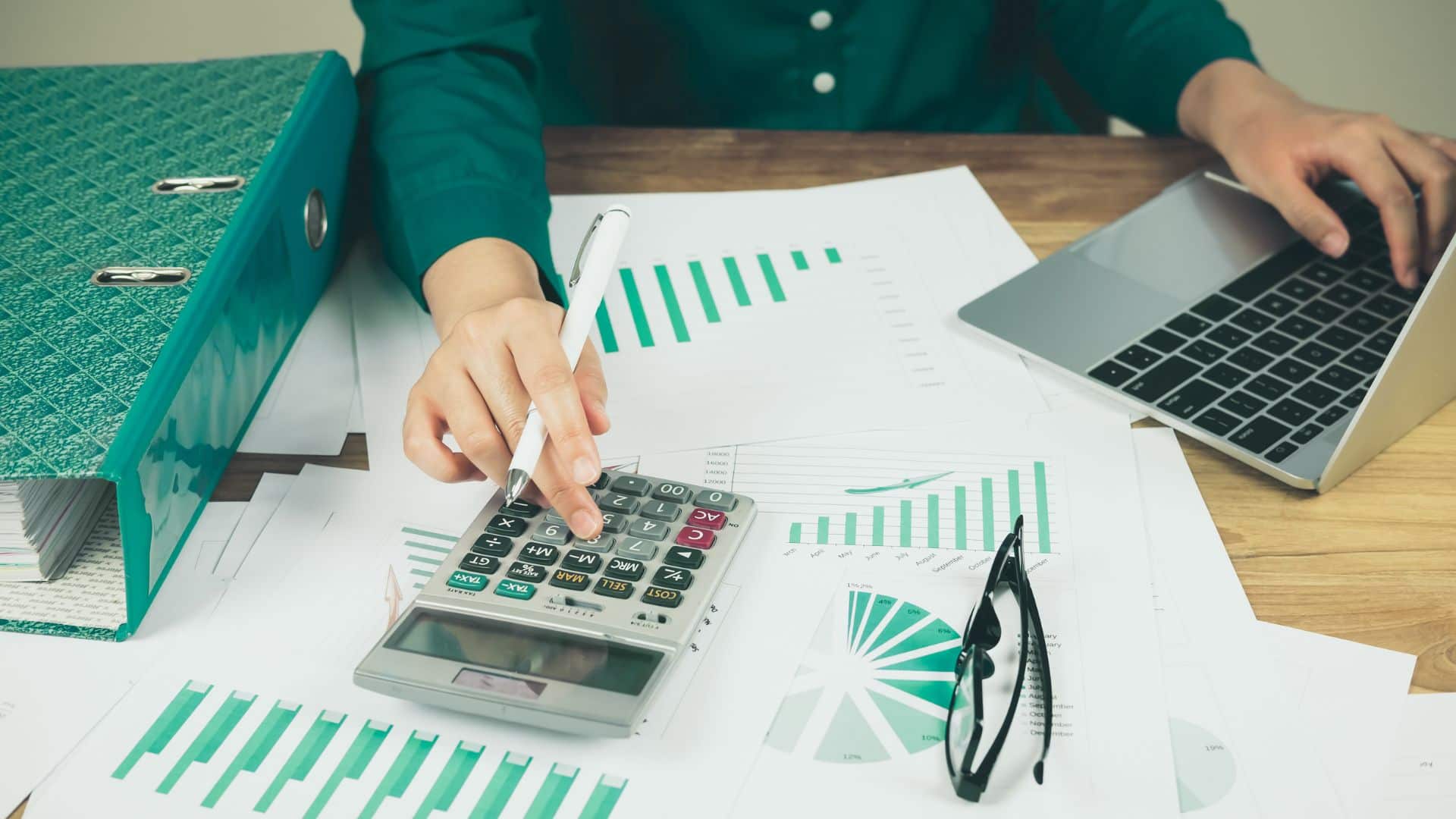 person pressing the keys of a calculator on their desk, they also hold a pen in the same hand, and have financial charts strewn over the surface, next to their laptop